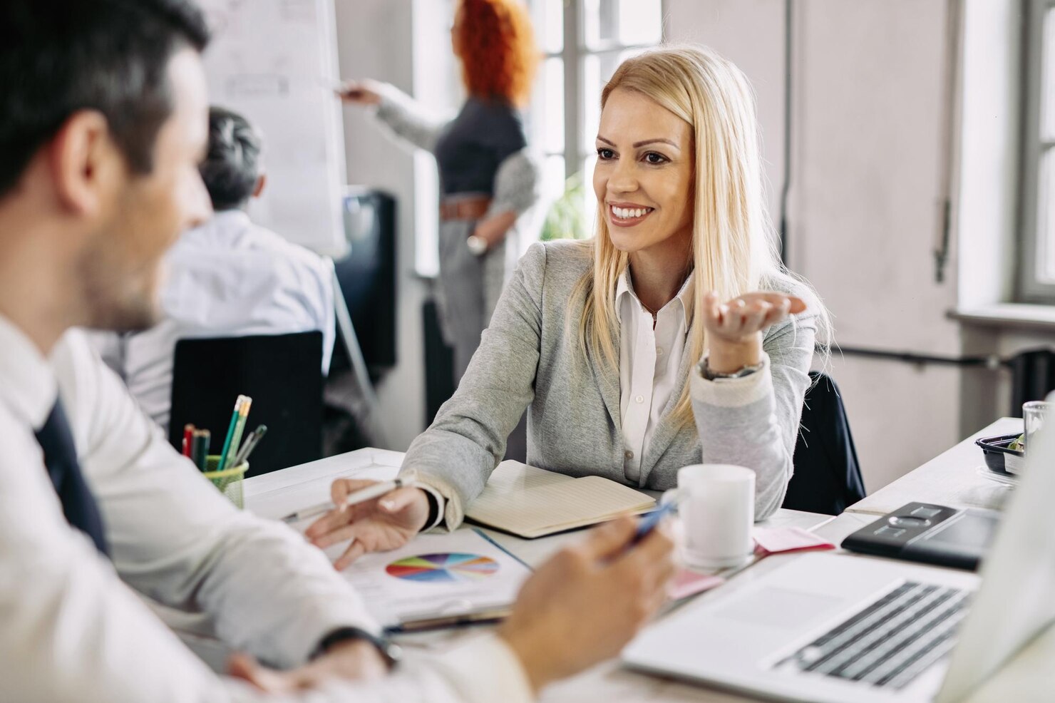 smiling-mid-adult-businesswoman-talking-developing-new-business-strategy-with-her-male-coworker-office-there-are-people-background_637285-259